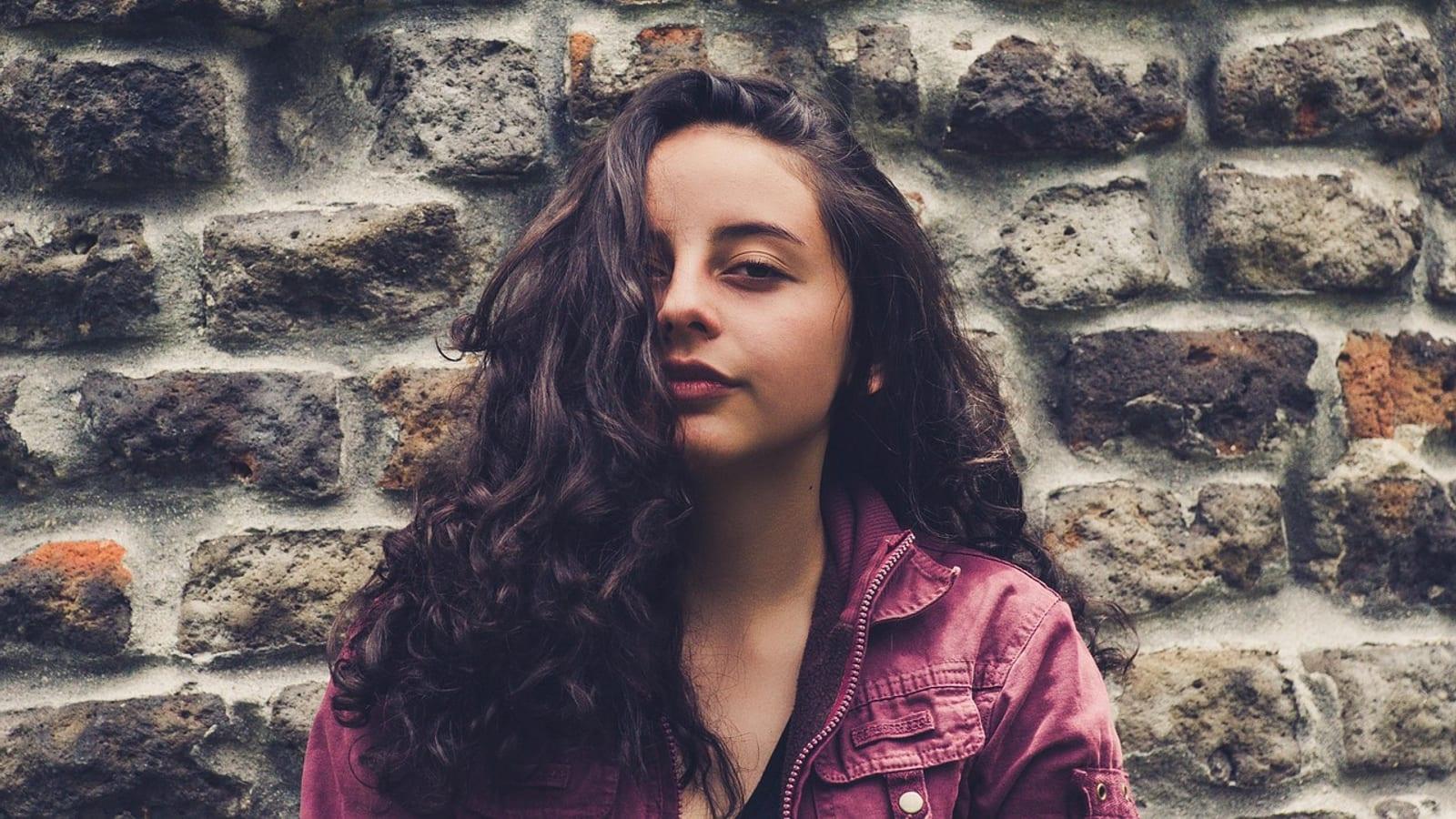 Young woman with long dark curly hair standing against a brick wall