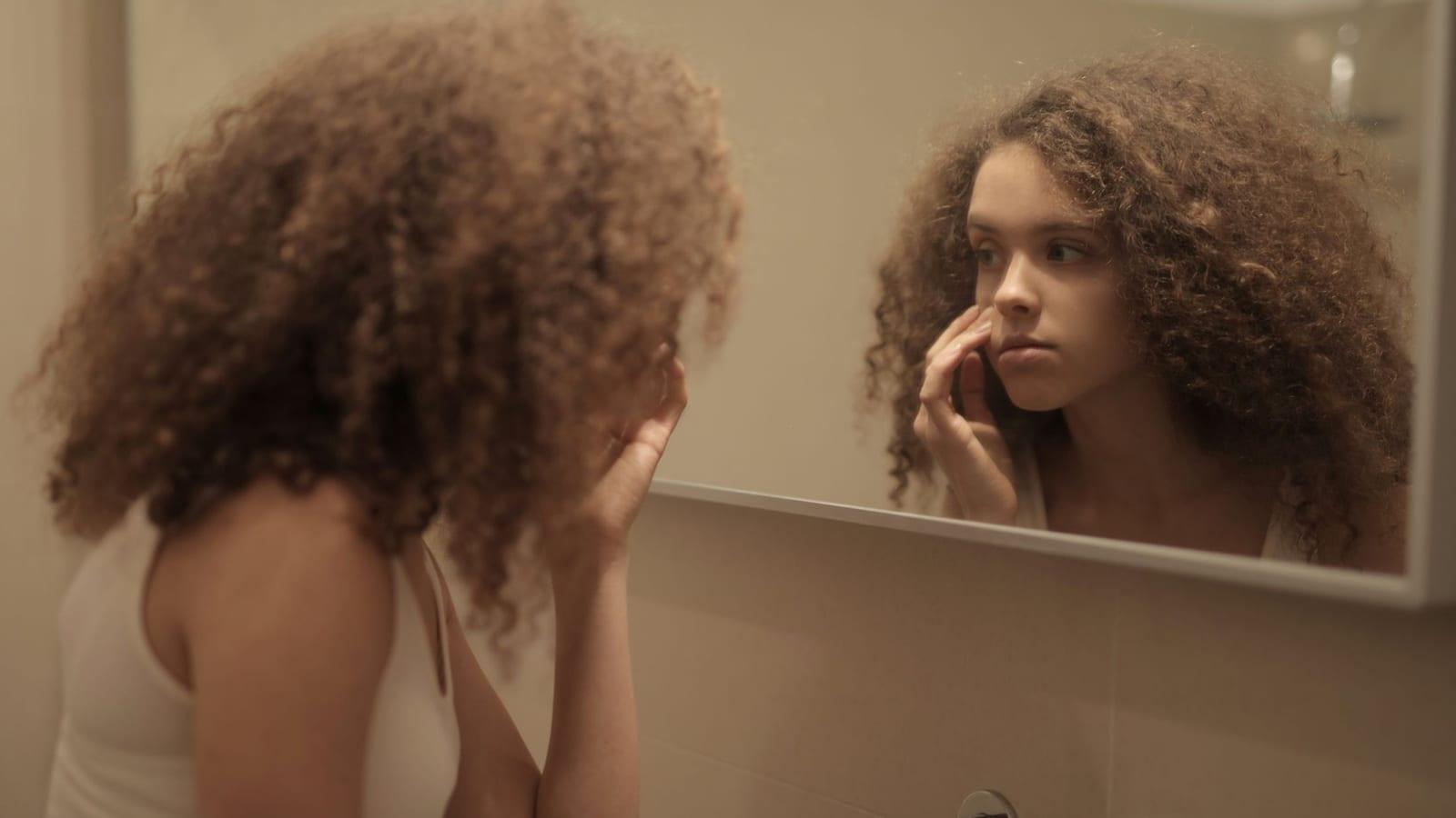Woman with big, frizzy curls studying her hair in a bathroom mirror