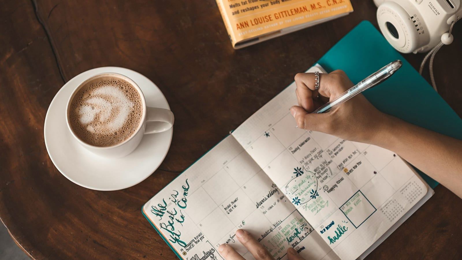 Hands writing in a planner next to a latte on a wooden table