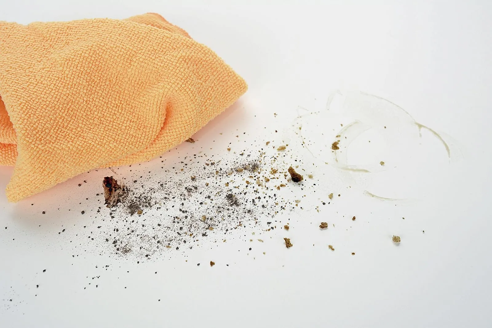 Orange microfiber cloth next to a scatter of crumbs and dust on a white surface