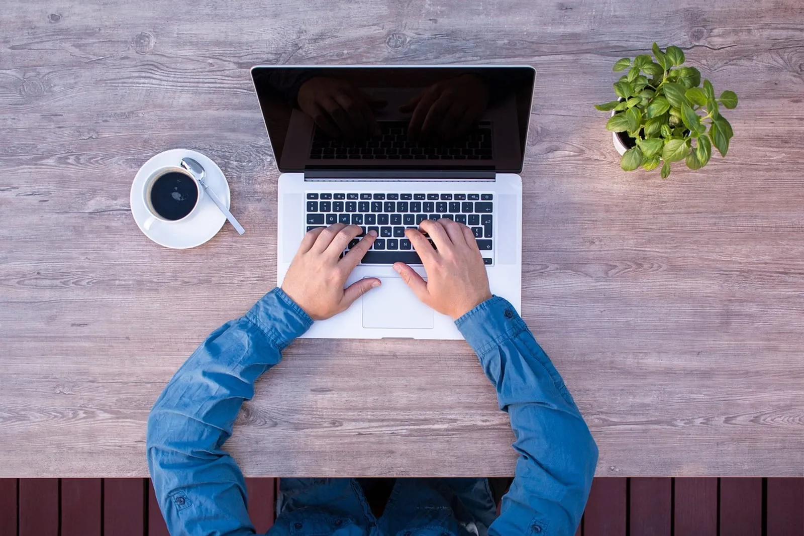 Person typing on a laptop at a wooden desk with a cup of coffee and a small plant, seen from above